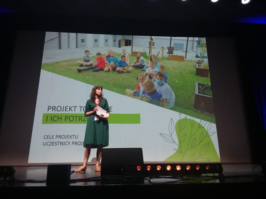 Zdjęcie z prezentacji na Ogólnopolskiej Giełdzie Projektów: "animacja + krajobraz". Projekt prezentuje w Agnieszka Michałowska ubrana w zieloną sukienkę. Za nią w tle wyświetlana jest prezentacja. Na niej między innymi śmiejące się dzieci w Zielonej Czytelni